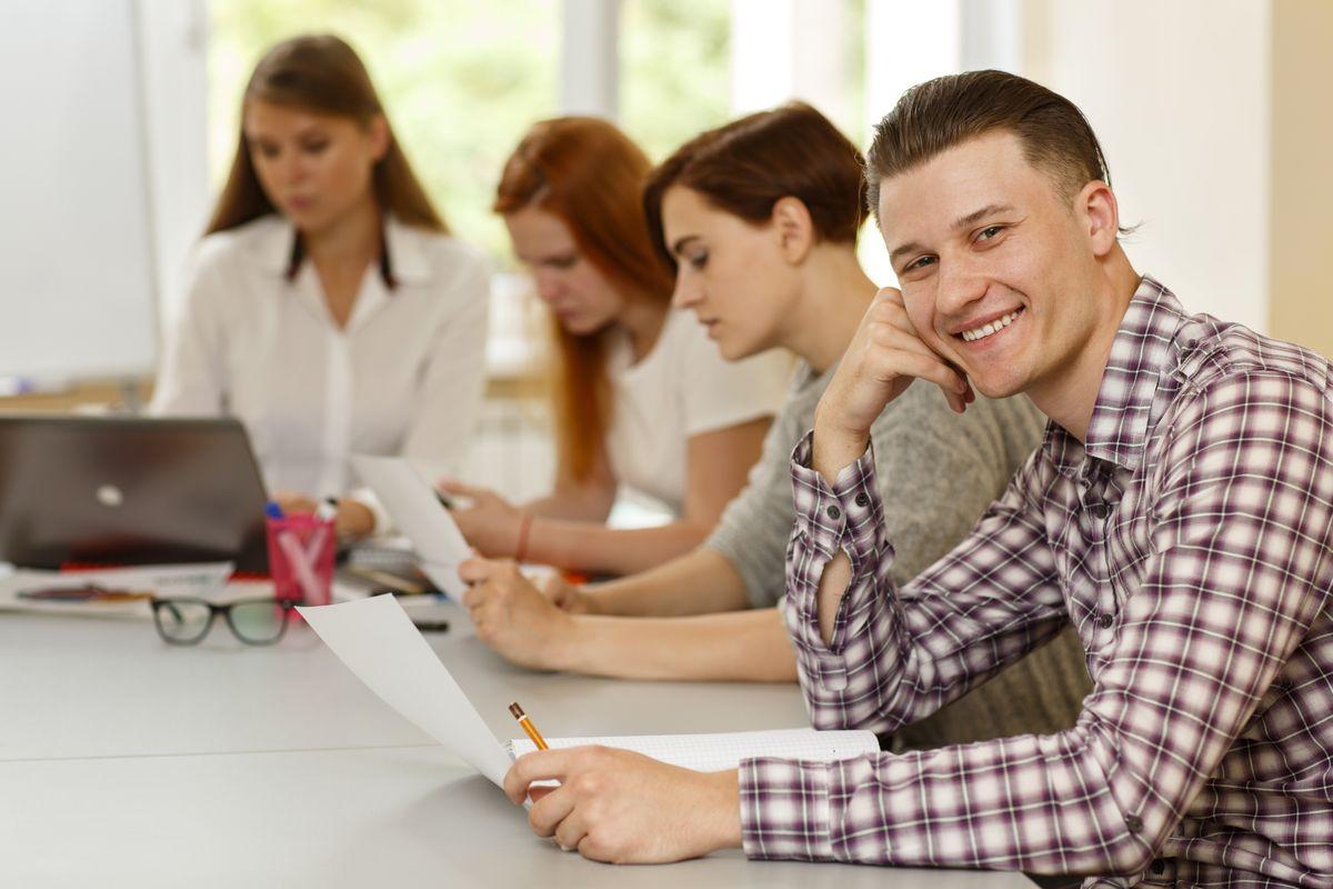 Positivity man sitting at table, looking at camera, leaning by hands and enjoying studying process. Clever female teacher helping and explaining material to students during lesson.