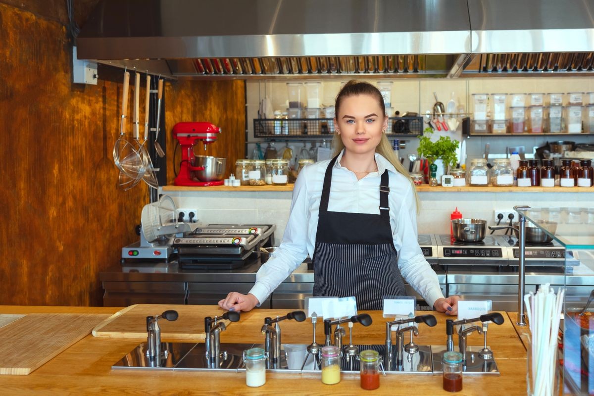 Smiling proud waitress woman standing behind counter looking at camera - Portrait of happy beautiful confident female wearing shirt with apron - Successful small business owner at restaurant eatery.